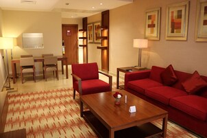 Habitación de hotel con sofá rojo, una mesita de centro de madera, un área de comedor con mesa y sillas, y un televisor.