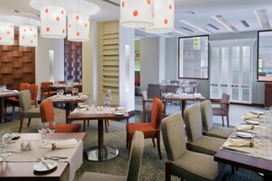 Comedor de hotel con varias mesas dispuestas para comer, sillas e iluminación colgante.