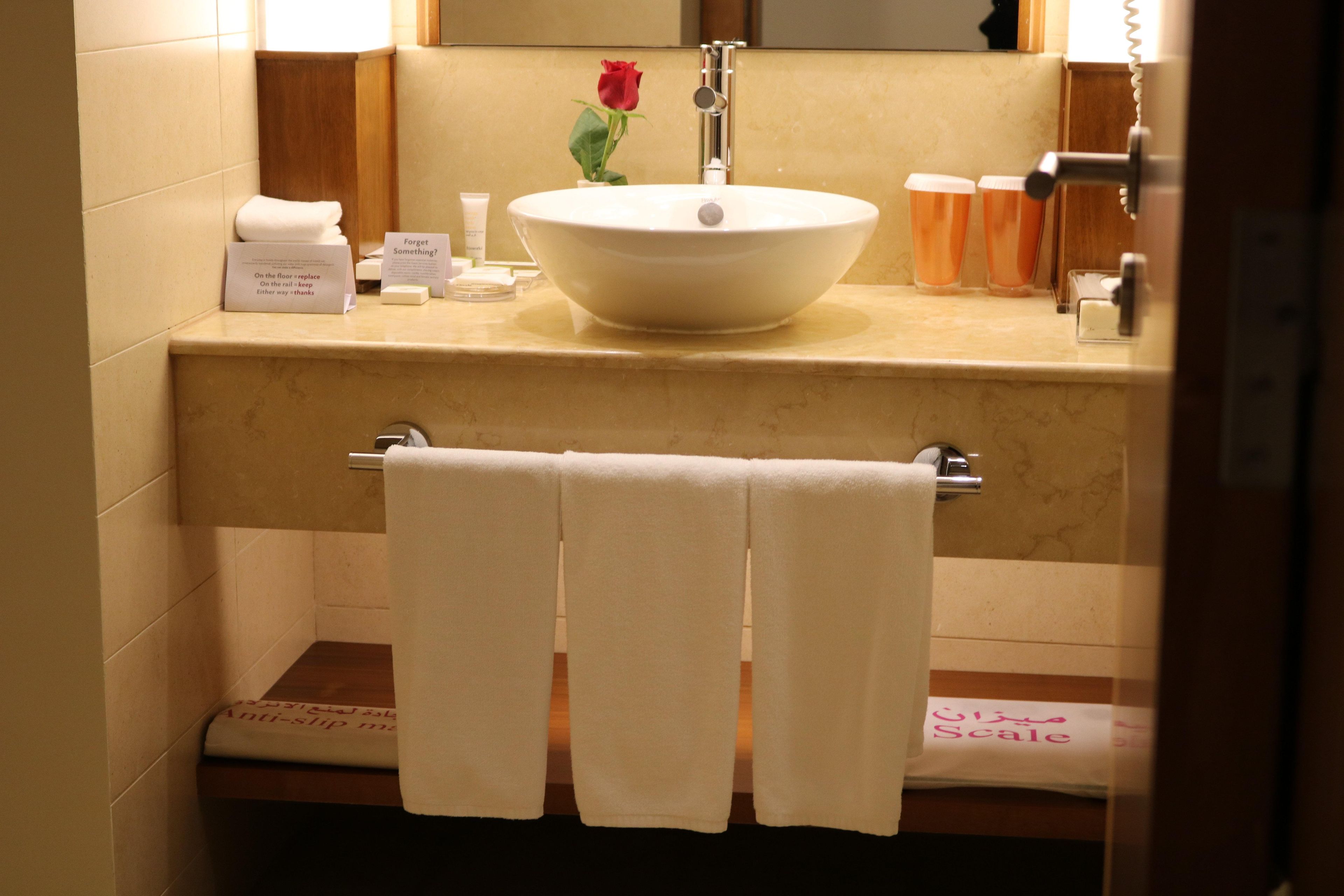 Un baño con un lavamanos blanco, un espejo, una rosa roja y un perchero para toallas.