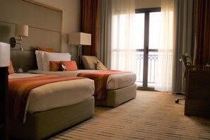 A hotel room with two beds, a desk, a chair, a lamp, and a view of the outside.