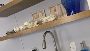 A kitchen with a stainless steel sink, wooden shelves with dishes, a blue vase, and a soap dispenser.