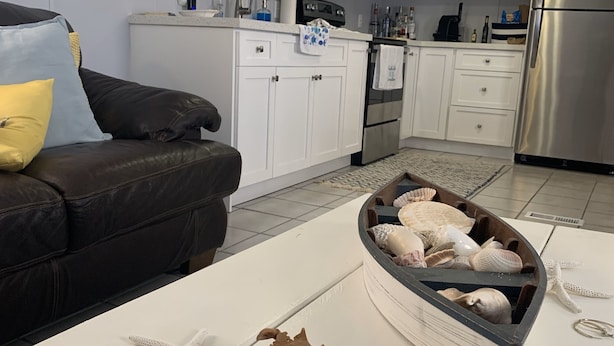 A modern kitchen with white cabinets, a stainless steel refrigerator, and a microwave. There is a wooden shelf above the sink with dishes and a blue vase. A brown leather couch is visible in the foreground.