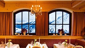 Uma sala de jantar com mesas preparadas para quatro pessoas, cada uma com um vaso de flores e uma vela. O espaço tem uma paleta de cores quentes em tons de vermelho e dourado, e grandes janelas com vista para montanhas cobertas de neve.
