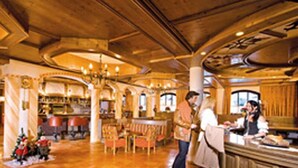 Um hall amplo com teto alto, vigas de madeira e um lustre grande. Há várias áreas de estar com mesas e cadeiras, e um tapete decorativo no chão.