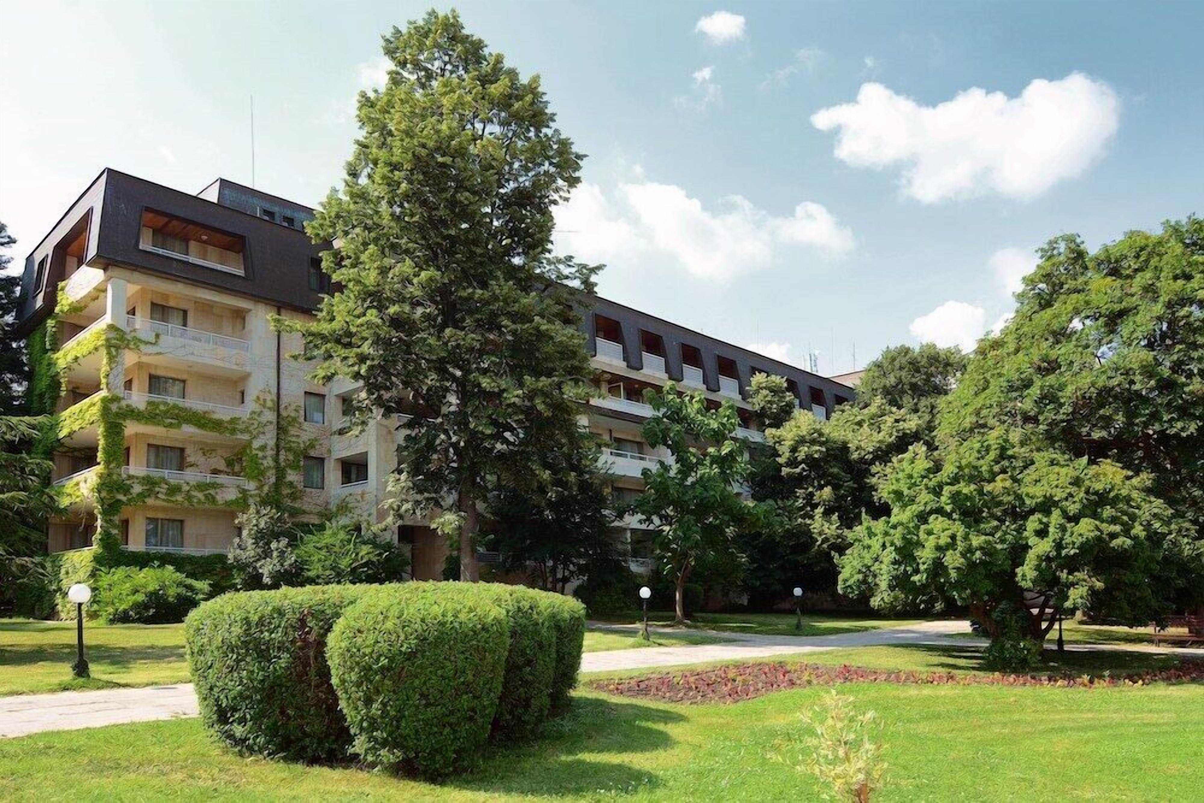 A multi-story building with a modern design, surrounded by well-maintained greenery and a clear sky.