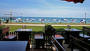 Un espace extérieur aménagé avec des tables et des bancs, offrant une vue sur une plage, des parasols et une jetée en arrière-plan.