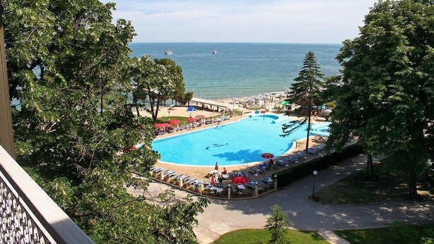 Un espace piscine doté de chaises longues et de parasols, entouré d’arbres et offrant une vue sur la mer.