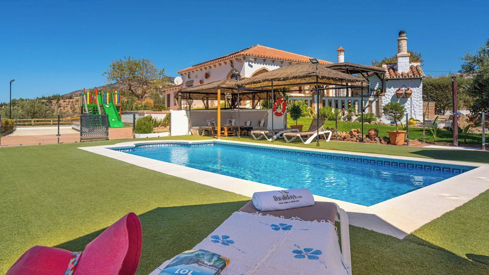 A swimming pool with lounge chairs, a covered seating area, and a building with a tiled roof.