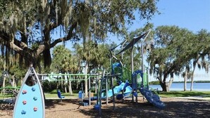 Un parque infantil con estructuras para escalar y toboganes, rodeado de árboles y un cuerpo de agua.