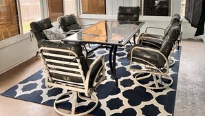 Un solárium con una mesa de vidrio rodeada por cuatro mecedoras, un tapete estampado y ventanas amplias.
