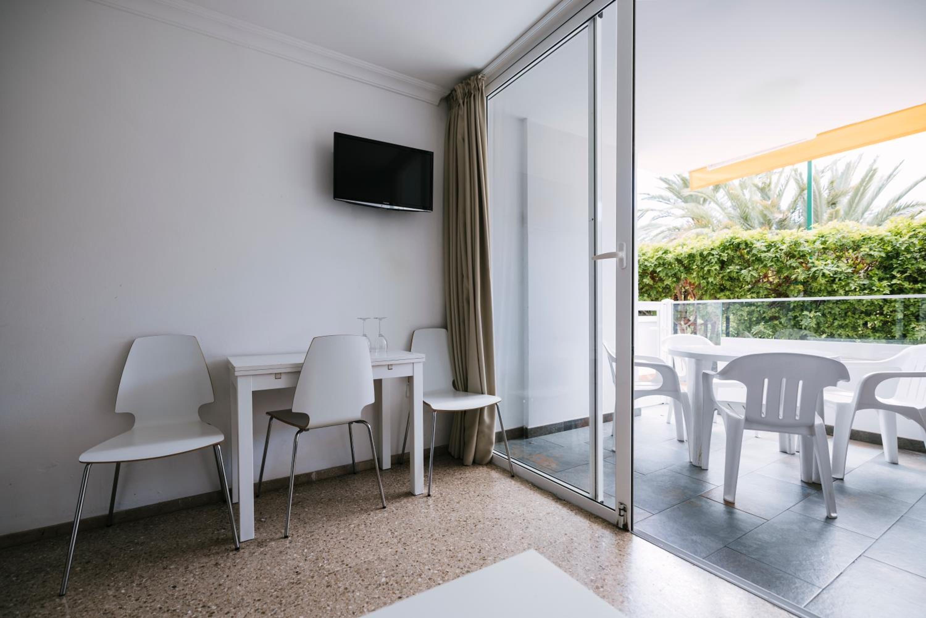 Une chambre moderne avec un bureau blanc, deux chaises, un téléviseur fixé au mur et une porte-fenêtre coulissante donnant sur un espace extérieur équipé de chaises en plastique blanches et d’un parasol jaune.