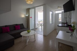 Un salon moderne avec un canapé, une petite table ronde et un coin cuisine équipé d’une télévision et agrémenté d’un vase de fleurs.