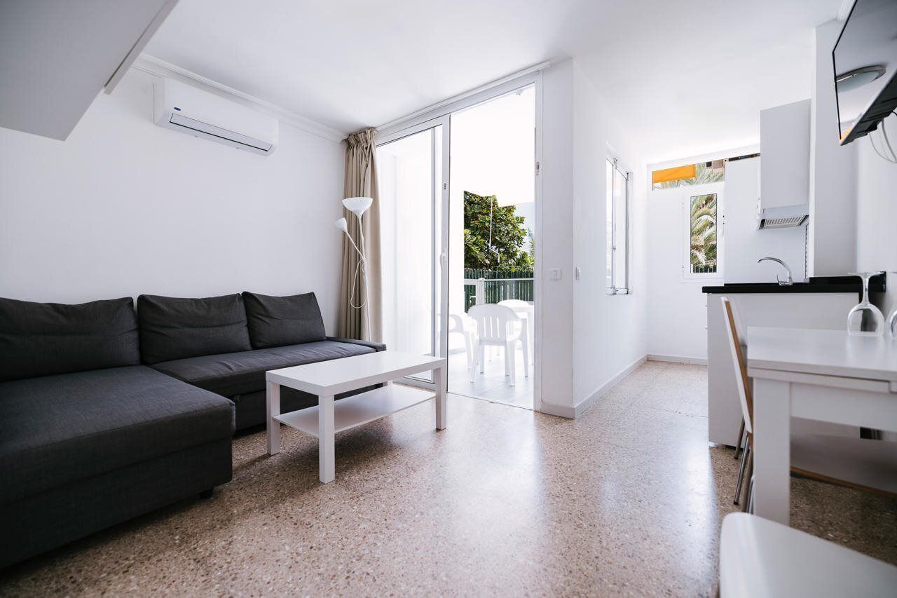 Una sala de estar moderna con un sofá gris, una mesa de centro blanca y una cocina con mostrador y lavamanos blancos.