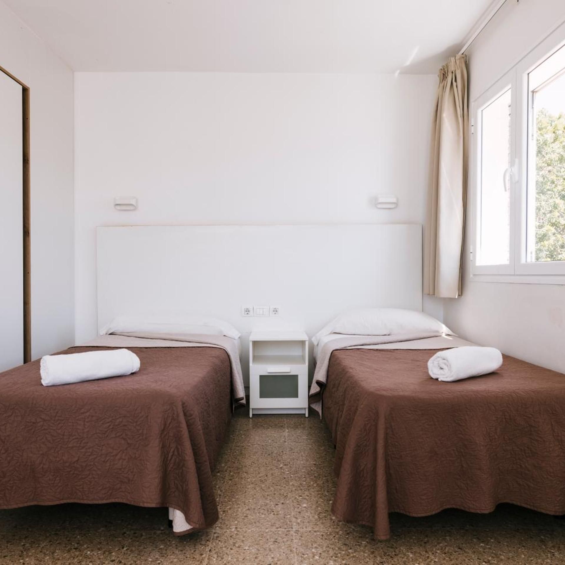A hotel room with two single beds, a small bedside table, a window with curtains, and a plain white wall.