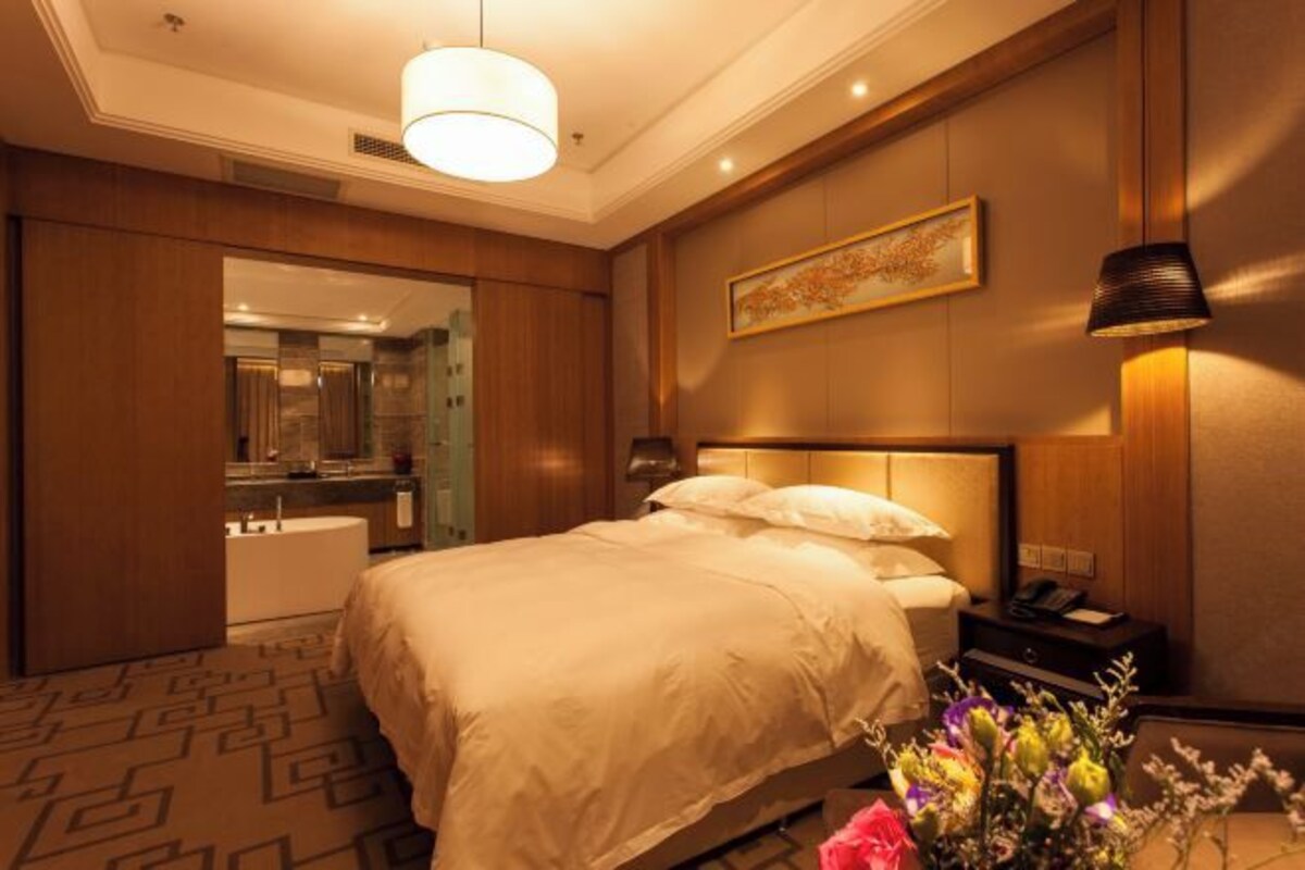 Una habitación de hotel con una cama grande, una mesita de noche, una lámpara, un cuadro en la pared y un baño visible a través de una puerta abierta.