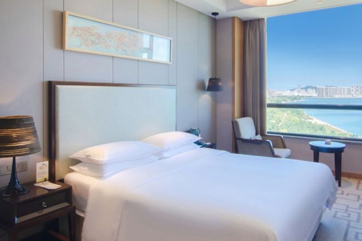 Habitación de hotel con una cama grande, un escritorio con lámpara, una silla y una ventana que ofrece vistas a un paisaje urbano y al agua.