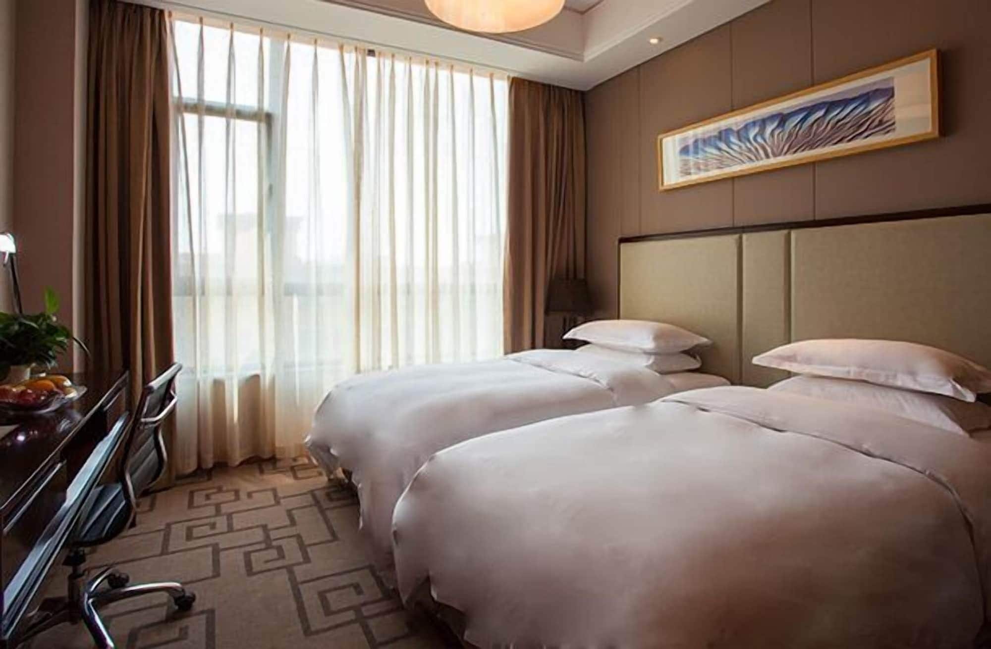 Une chambre d’hôtel avec deux lits, un bureau avec une chaise, une grande fenêtre avec des rideaux et une œuvre d’art encadrée au mur.