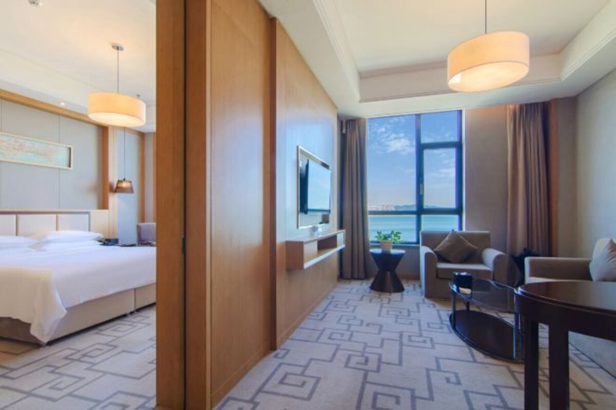 Habitación de hotel con cama, televisor, sofá, mesa de comedor y una ventana grande con vistas.