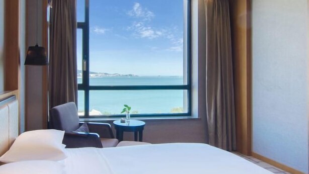 A hotel room with a large window offering a sea view, a bed with white linens, a small round table with a plant, and a chair. - Guihe Siji Hotel Weihai (Weihai)