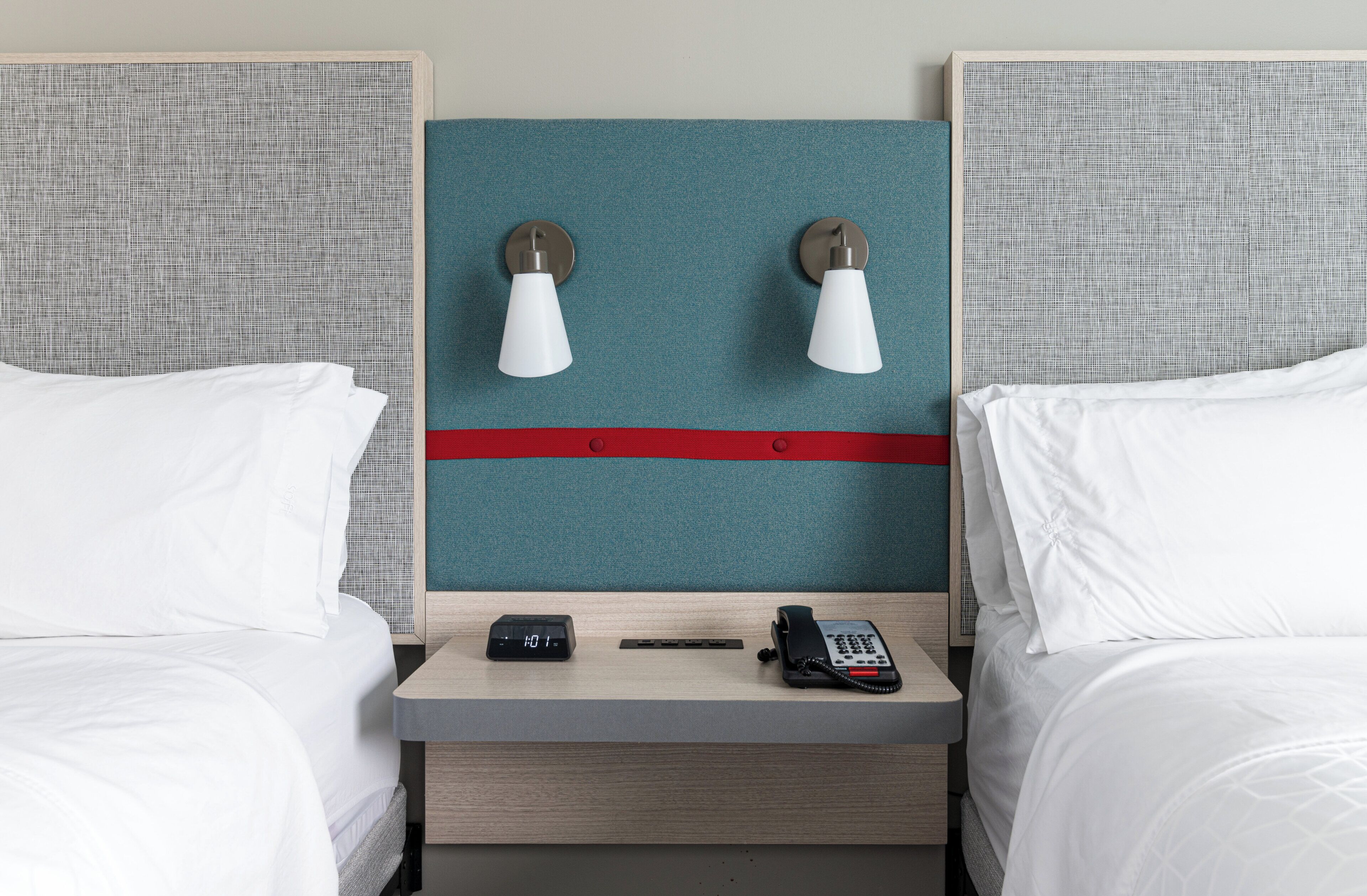 Een hotelkamer met twee bedden, een nachtkastje, een digitale klok, een telefoon en wandlampen.