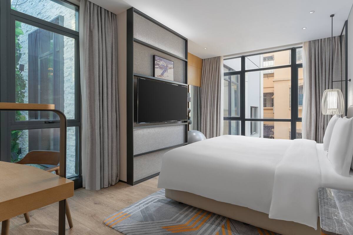 A modern hotel room with a large bed, a desk, a TV, and a view of the city.