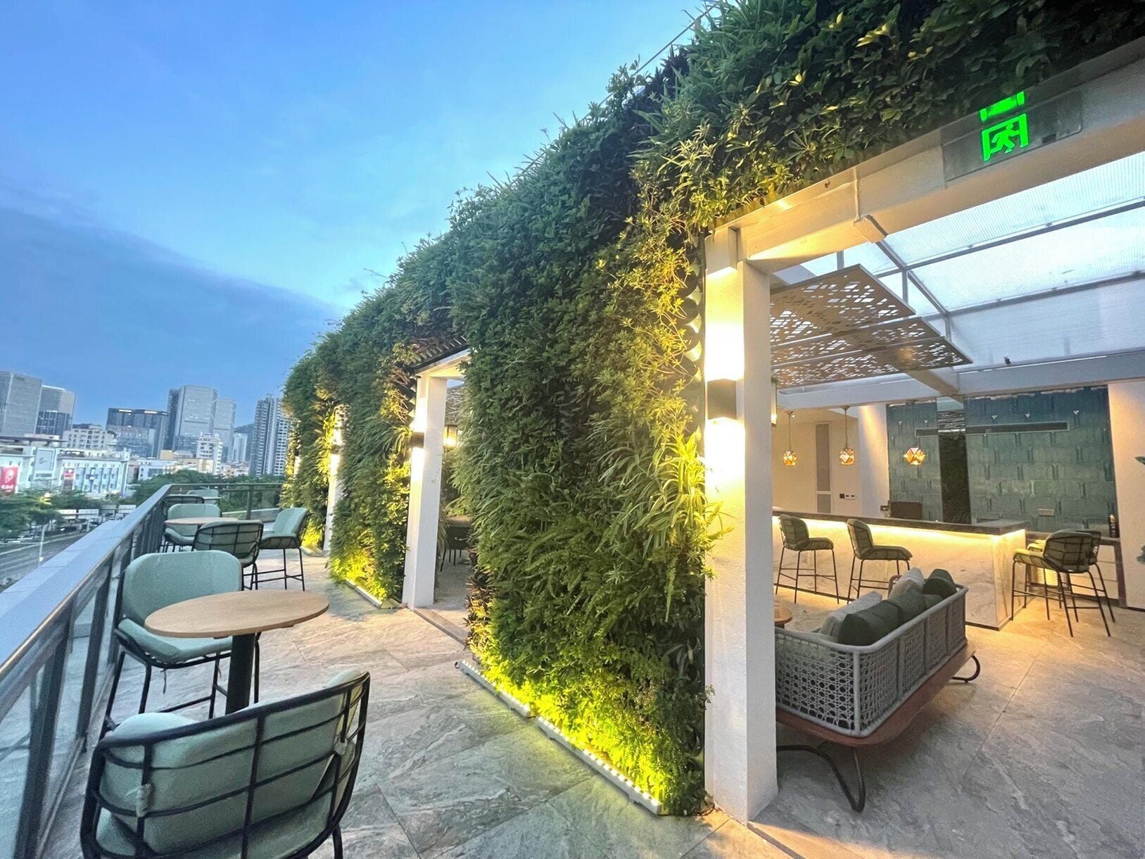An outdoor seating area with tables and chairs, a green wall with plants, and a city skyline in the background.