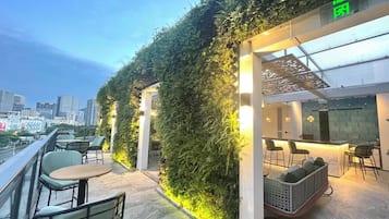 An outdoor seating area with tables and chairs, a green wall with plants, and a city skyline in the background.