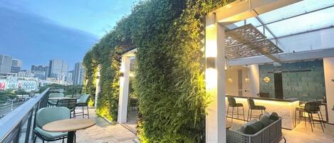 An outdoor seating area with tables and chairs, a green wall with plants, and a city skyline in the background.