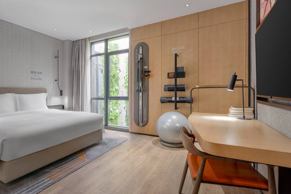 A modern hotel room with a large bed, a desk with a chair, a standing exercise machine, and a large window offering a view of greenery.