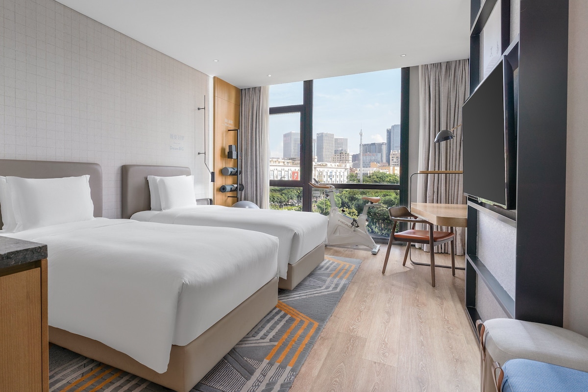 A hotel room with two beds, a desk, a chair, a TV, and a city view.