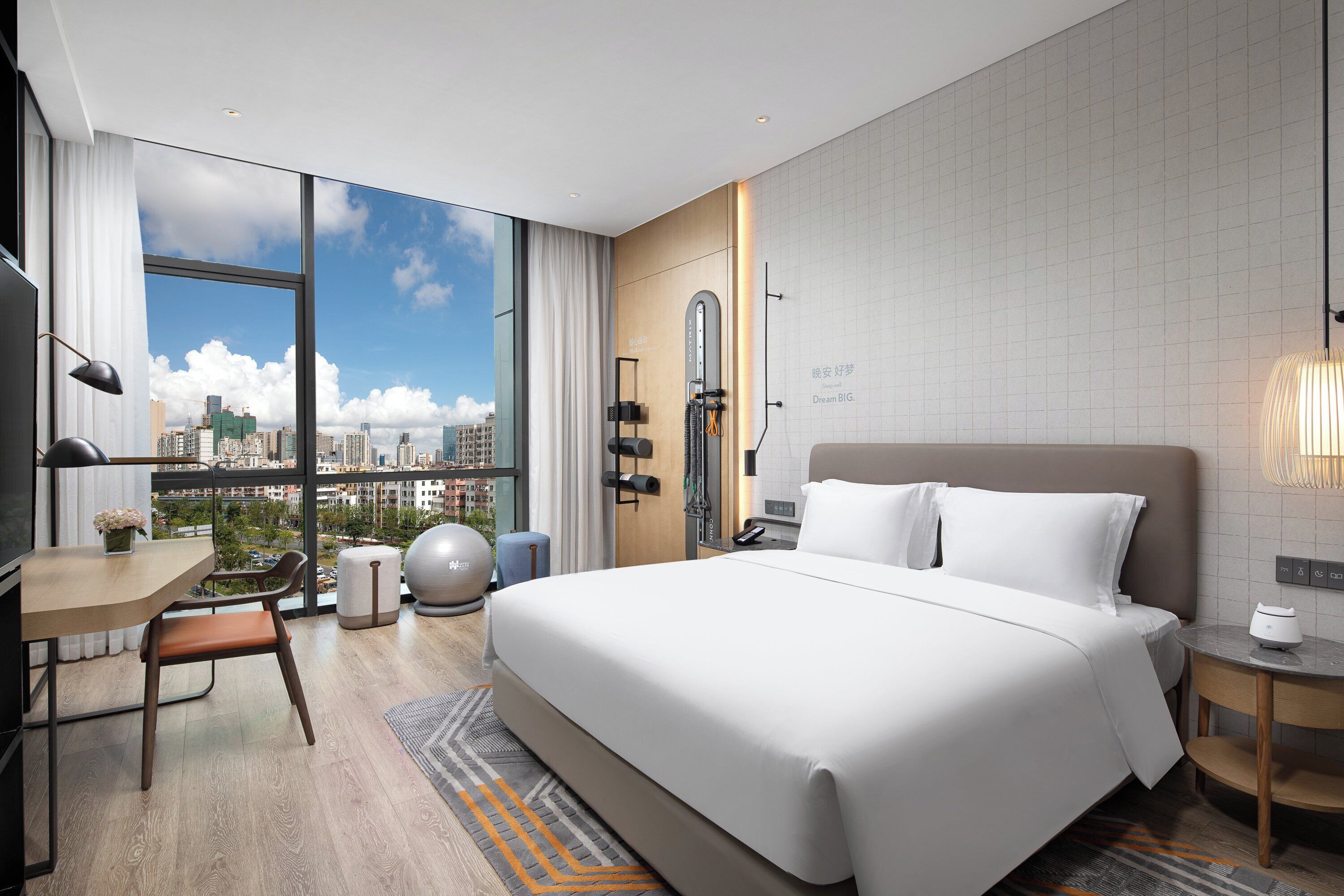 A modern hotel room with a large bed, a desk, and a view of the city skyline through floor-to-ceiling windows.