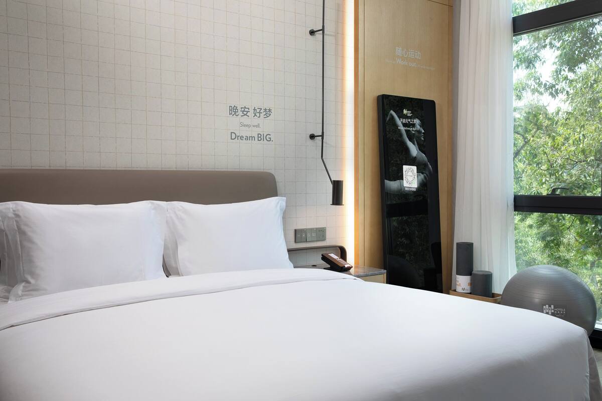 A hotel room with a neatly made bed, a wall-mounted TV, a window with a view of trees, and a small exercise ball.