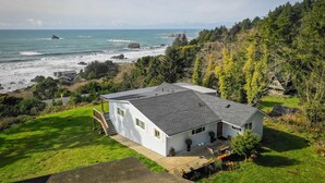 A single-story house with a grey roof, situated on a grassy hillside overlooking a rocky coastline. - Beautiful Ocean View Home on the Majestic Oregon Coast The Blue Pelican (Brookings)