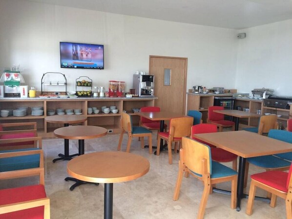 Een eetgedeelte met ronde tafels en stoelen, een koffieapparaat en een aan de muur bevestigde televisie.