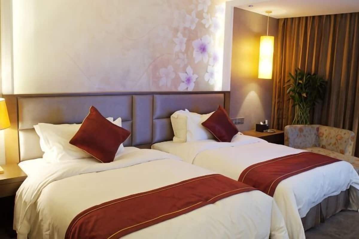 A hotel room with two beds, a nightstand, a lamp, a chair, and a large window with curtains.