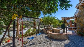 Un parque infantil con columpios y una estructura para escalar, rodeado de árboles y un edificio de piedra.