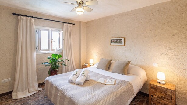 A neatly arranged bedroom with a bed, bedside lamps, a window with curtains, a plant, and a framed picture on the wall. - El Balcón del Olivar Villanueva de Algaidas by Ruralidays (Málaga)