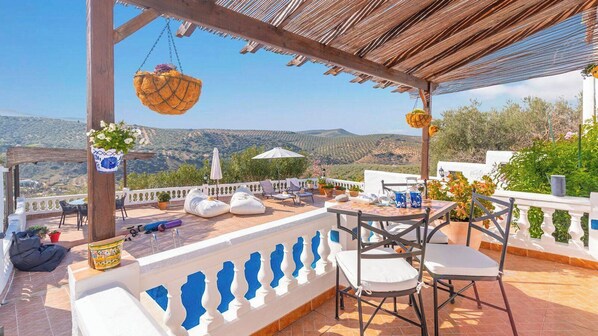 Terraza al aire libre con vistas al campo, amueblada con sillas, una mesa y macetas colgantes.