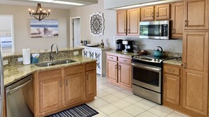 Une cuisine moderne dotée de plans de travail en granit, d’armoires en bois, d’appareils électroménagers en acier inoxydable et offrant une vue sur la plage grâce à une grande fenêtre.
