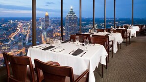 Un restaurante con mesas dispuestas para comer, ventanas amplias que ofrecen vistas de la ciudad y un techo alto.