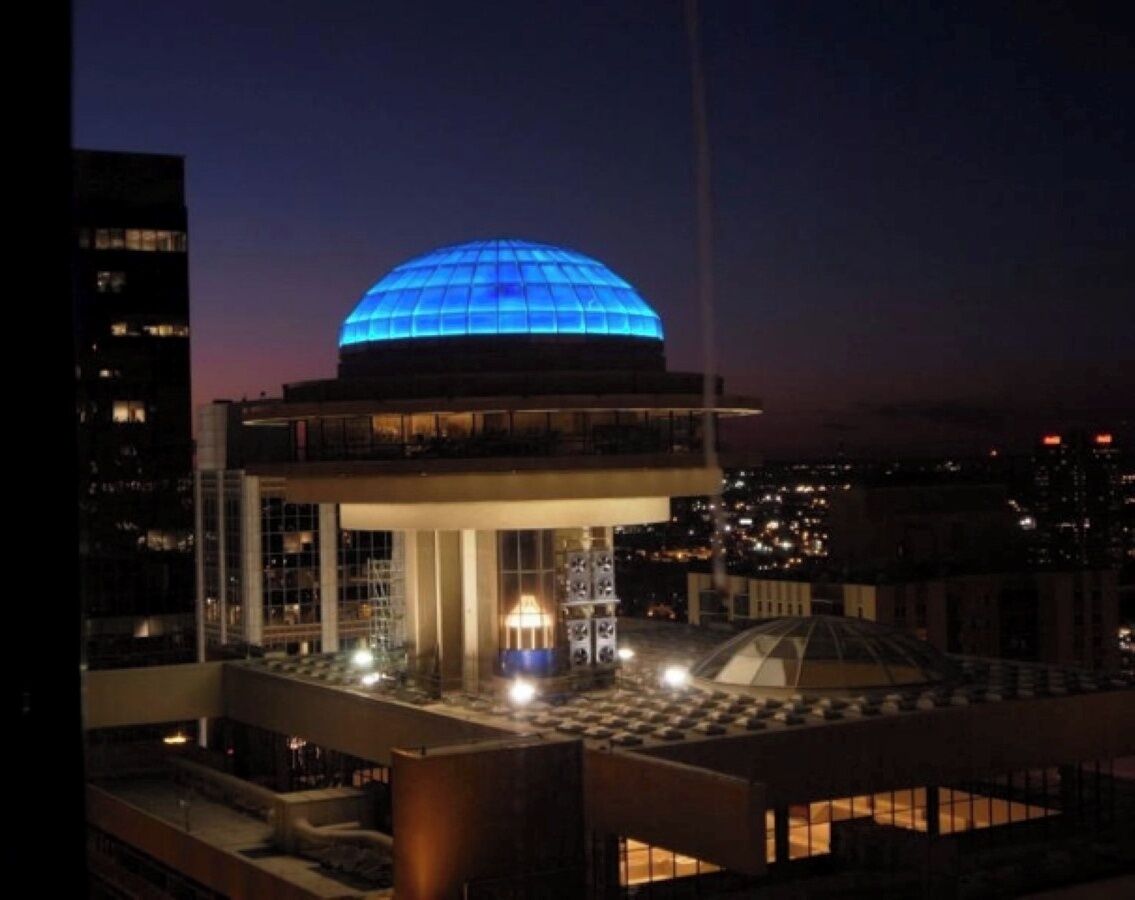 A modern building with a large blue dome at the top, surrounded by a lower level with a glass facade and a central light fixture.