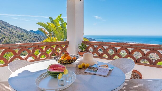 Une table avec un saladier de fruits, une tranche de pastèque et un chapeau, sur une terrasse offrant une vue sur la mer et les montagnes.