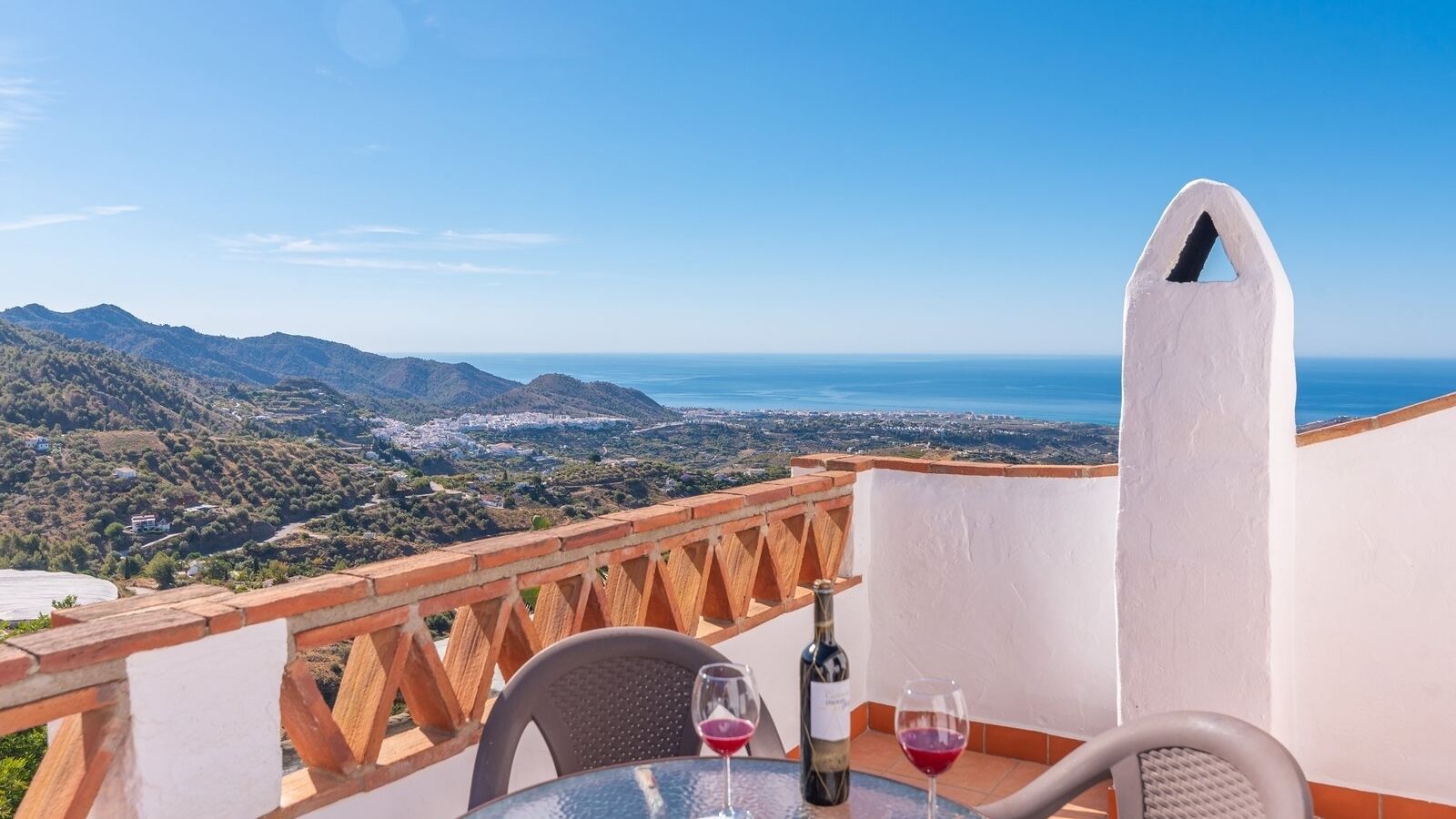 A terrace with a table, two wine glasses, and a bottle of wine. The terrace overlooks a mountainous landscape and the sea.