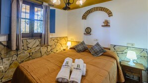 A hotel room with a stone wall, a bed with a brown bedspread, two towels with the hotel logo, a nightstand with a lamp, and a window with curtains. - Villa Javier de Frigiliana by Ruralidays (Málaga)