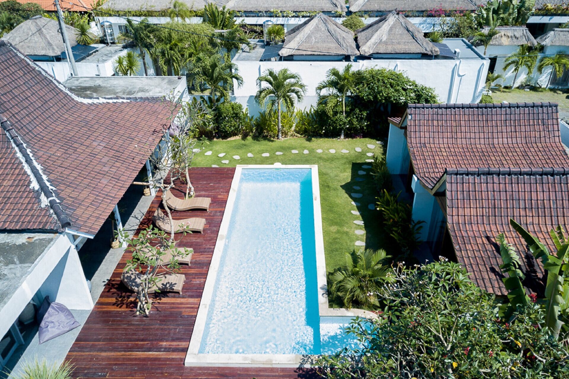 A rectangular swimming pool with a wooden deck and lounge chairs, surrounded by tropical vegetation and thatched-roof buildings.