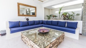 A modern living room with a blue sectional sofa, a glass coffee table with a natural wood pattern, a large mirror, and a view of a balcony with plants.