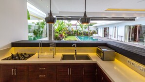 A modern kitchen with a built-in island, two pendant lights, a view of a swimming pool, and a glass door leading to a covered outdoor area. - 5 Star Private Villa Holiday in Seminyak at an Unbeatable Rate, Bali Villa 1158 (Bali)
