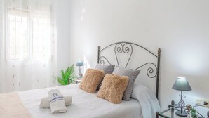 Una cama bien tendida con almohadillas mullidas, una mesita de noche con lámpara y una ventana con cortinas translúcidas.