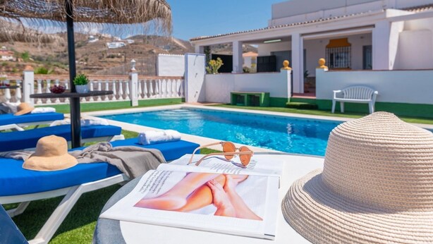 Zona de piscina con sillones de descanso, piscina y edificio de fachada blanca.