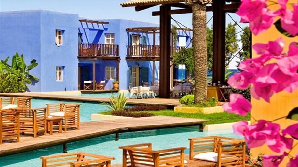 A pool area with wooden loungers and a blue building with balconies. - Miramare Park (Rhodes)